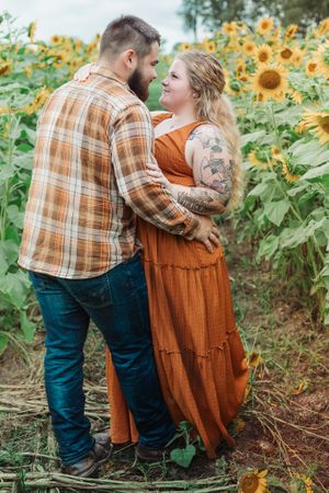 A couple embraces tenderly among towering sunflowers, with visible tattoos and rustic autumn clothing.