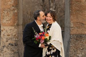 A couple shares an intimate moment with a vibrant bouquet against stone columns.