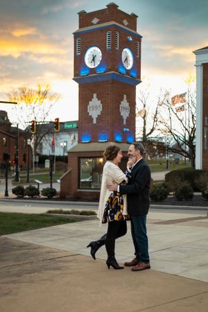 A couple embraces in front of an illuminated clock tower at sunset.