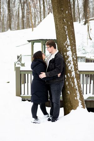 A couple embraces near a large tree trunk on a snowy winter deck with white-covered woods in the background.