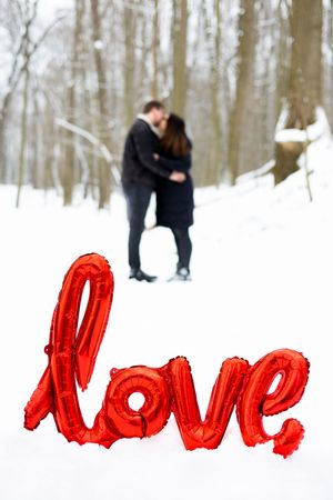 A romantic couple embraces in a snowy winter forest scene with bright red text overlaid on the white snow.