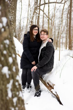 A couple in winter attire playfully poses in the snow against a backdrop of bare trees in the forest.