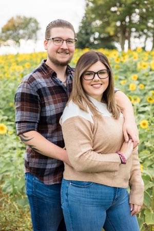 A couple stands together smiling in a field of bright yellow sunflowers on a sunny autumn day.