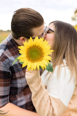 A couple embraces behind a vibrant yellow sunflower in outdoor photos showing romantic moments during a photoshoot.