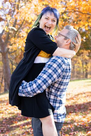 A couple shares a playful embrace while laughing together among vibrant autumn foliage in a park setting.