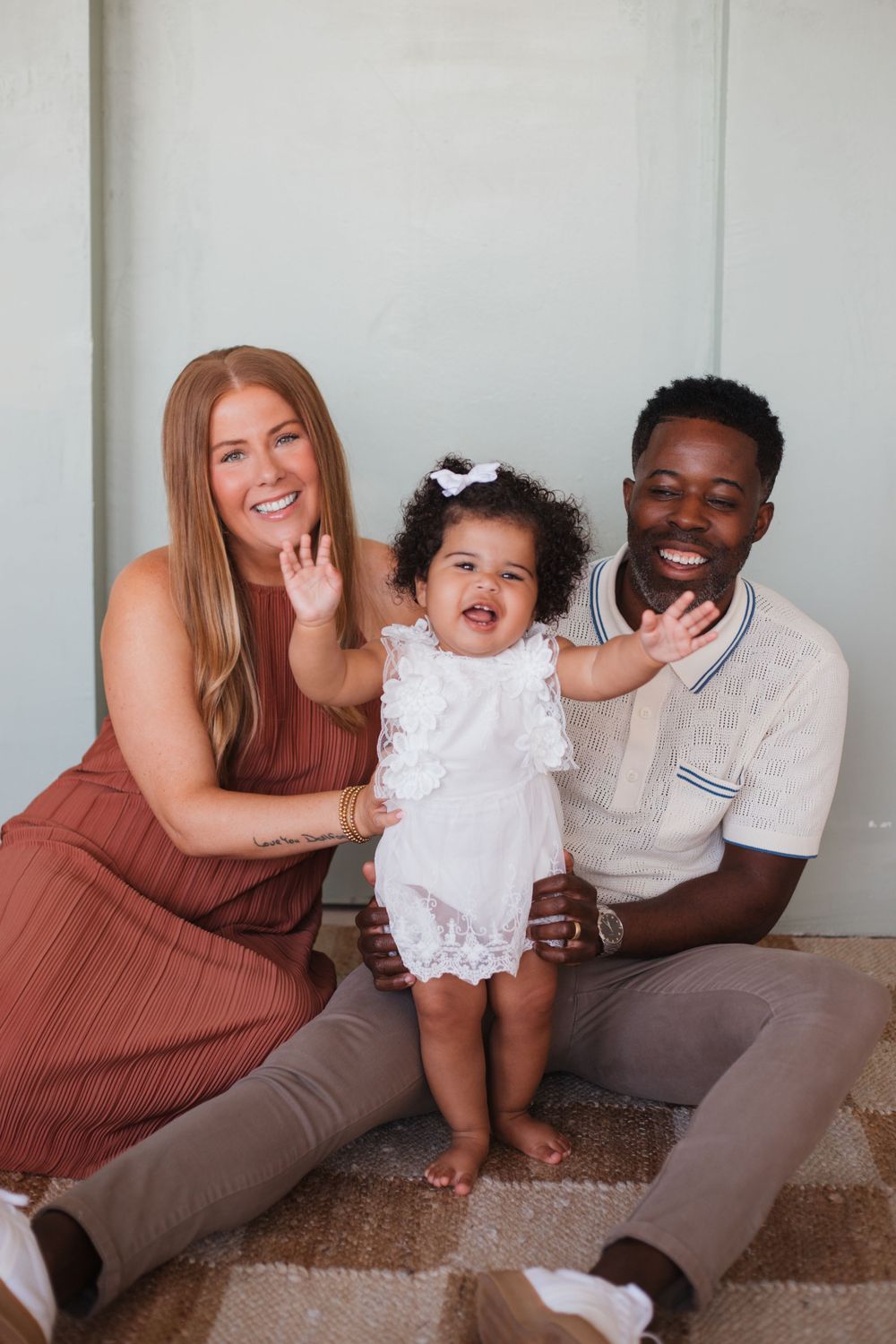 Interracial family with young child in white dress poses together on carpet for casual indoor photoshoot.