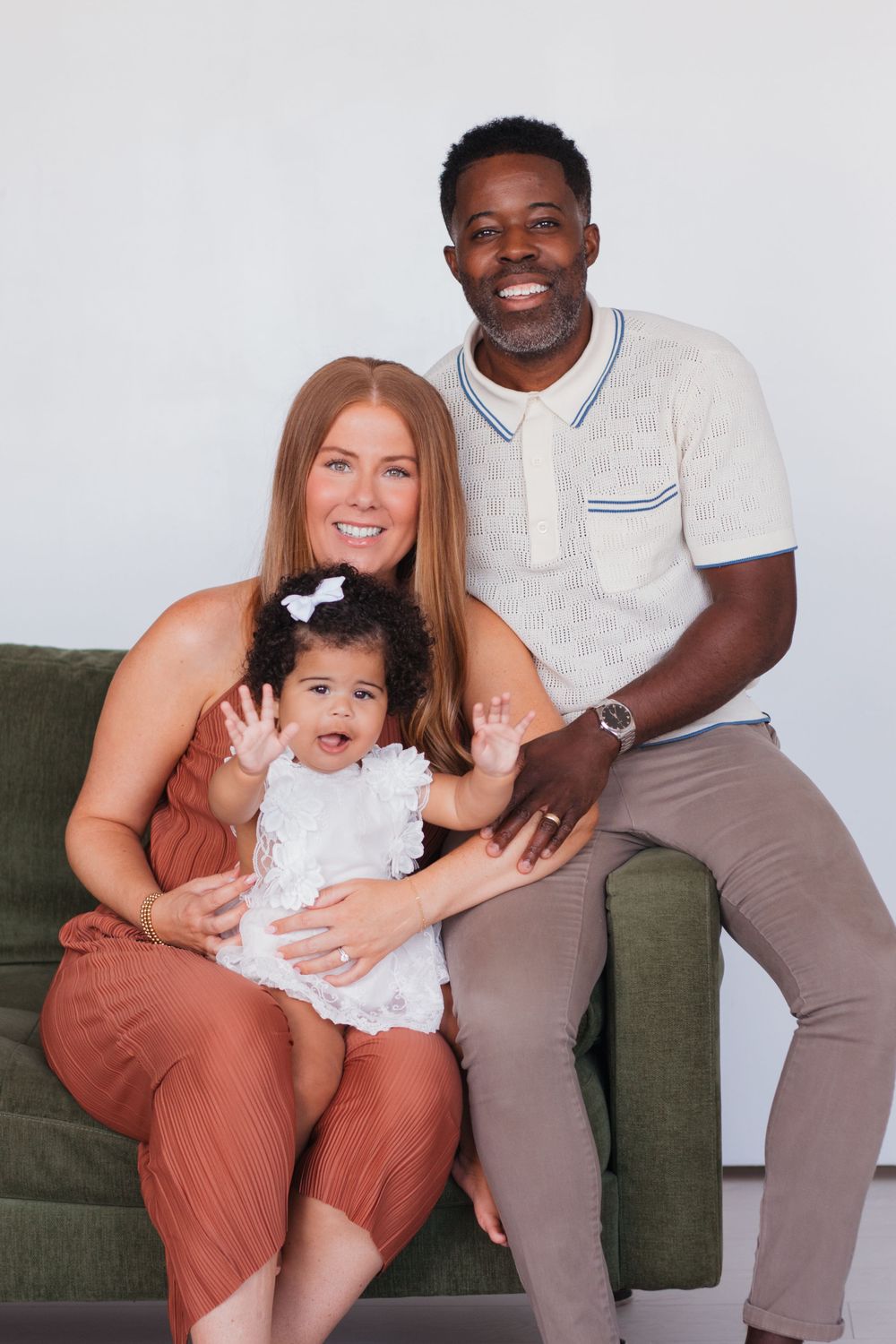 Family of three poses on green couch for studio portrait in coordinated casual outfits.