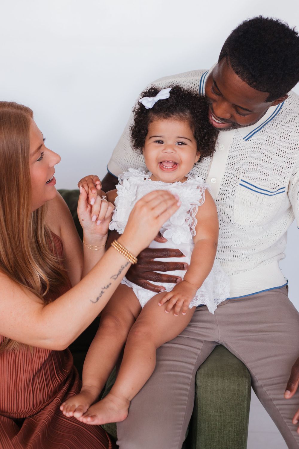 Happy family shares joyful moment while holding laughing toddler in white dress.