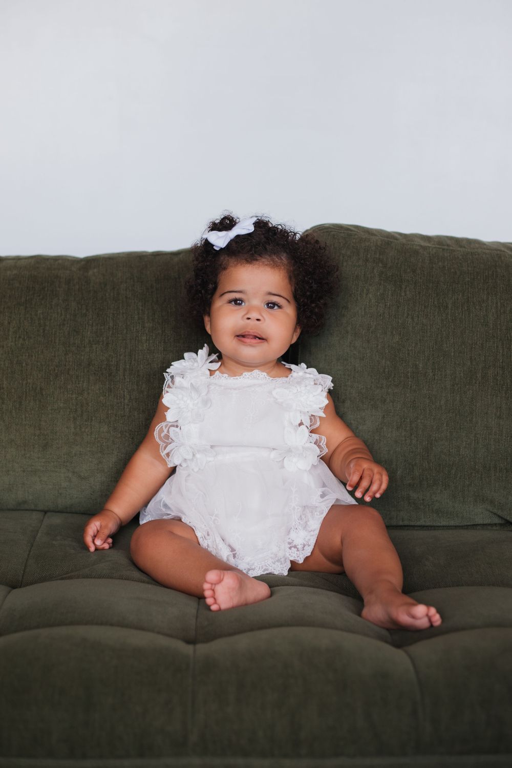 Adorable toddler in white lace outfit sits smiling on green couch.