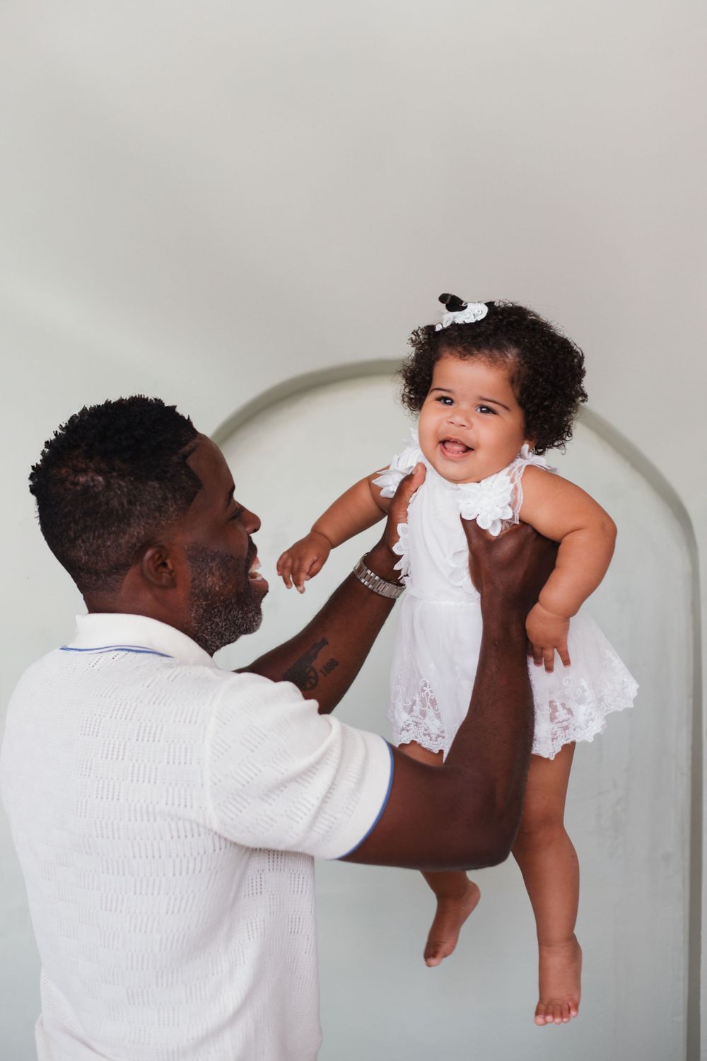 A joyful moment captured as a child in white lace dress is lifted up against an arched backdrop.