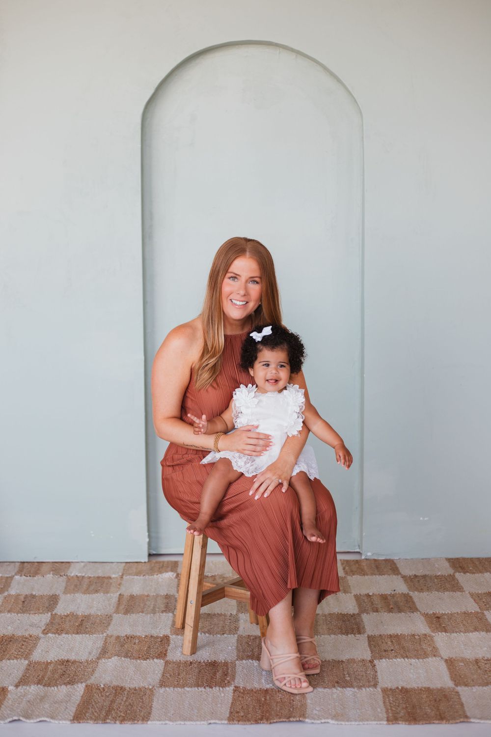 A woman and child sit together on a wooden stool in front of an arched white wall.