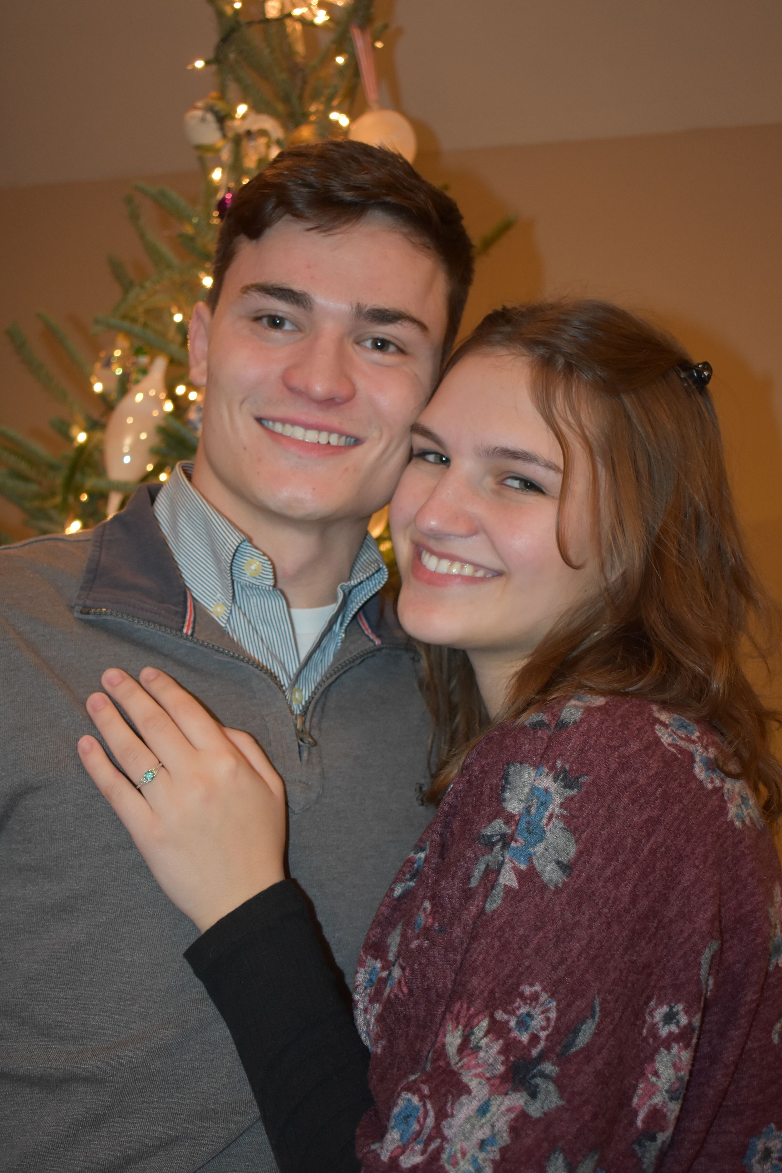 Newly engaged couple smiling together in front of illuminated Christmas tree.