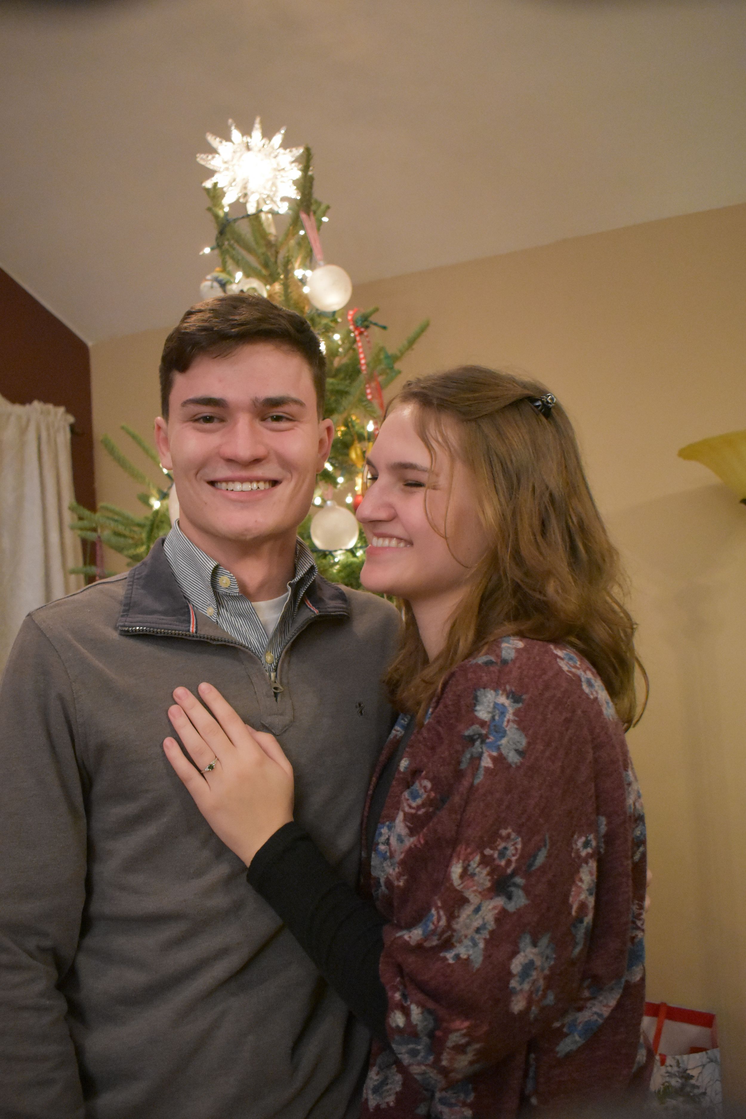 Happy couple poses in front of lit Christmas tree during engagement celebration.