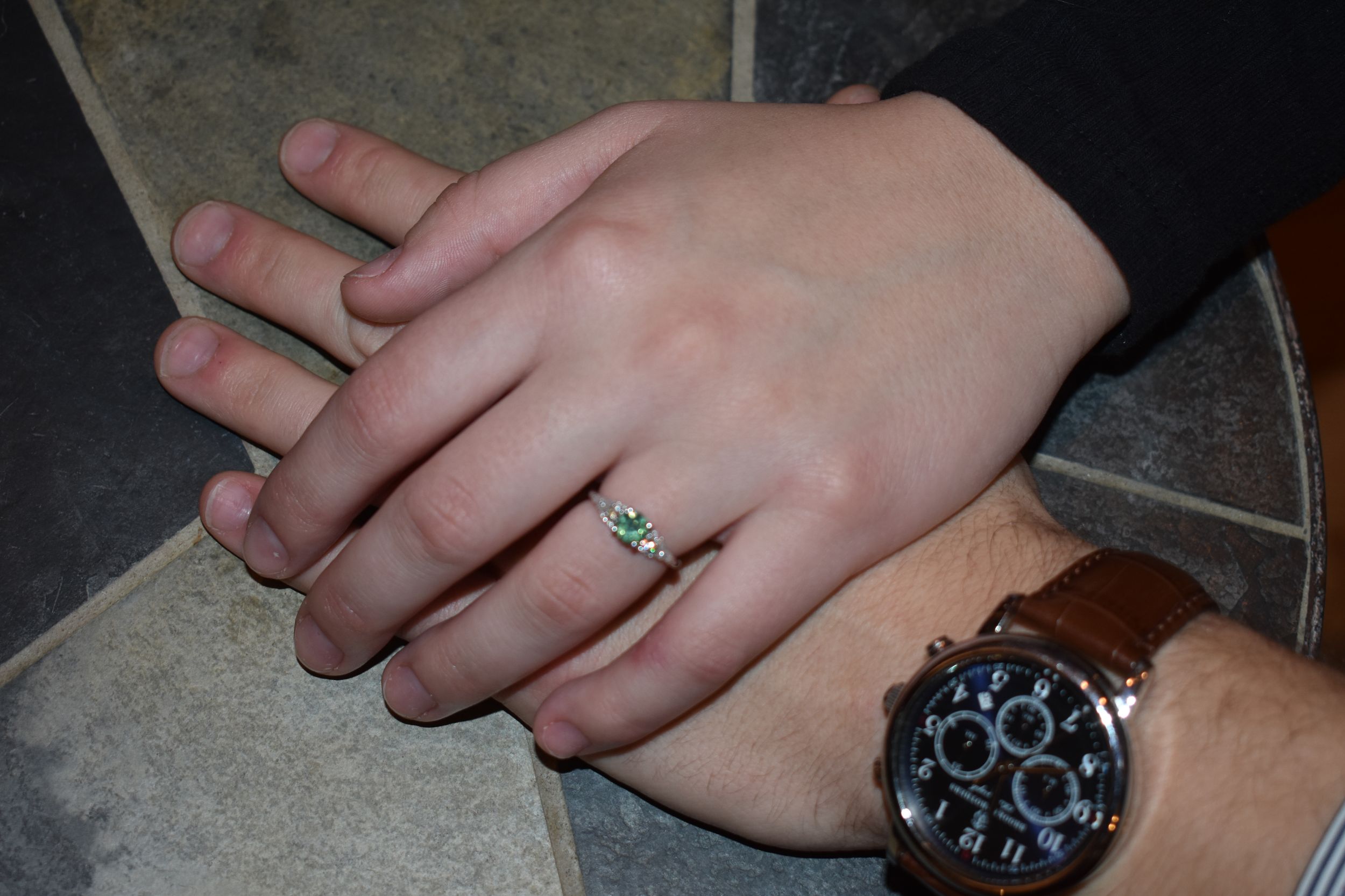 Engaged couple's hands showing off ring and watch on gray surface.