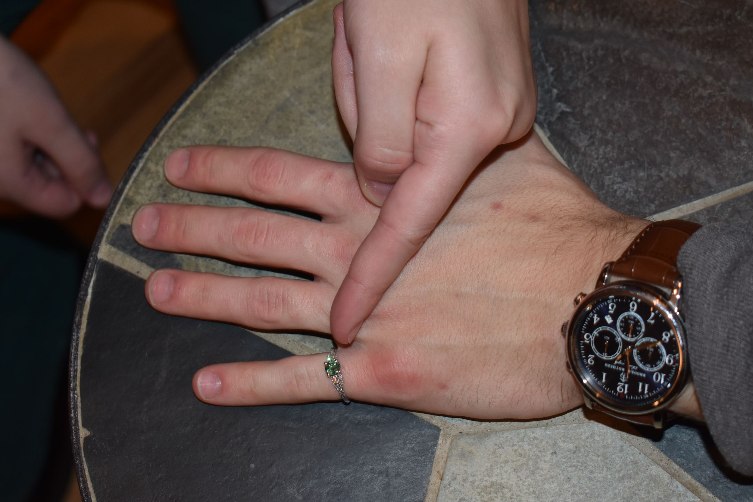 Close-up of hands displaying engagement ring and watch on a gray surface.