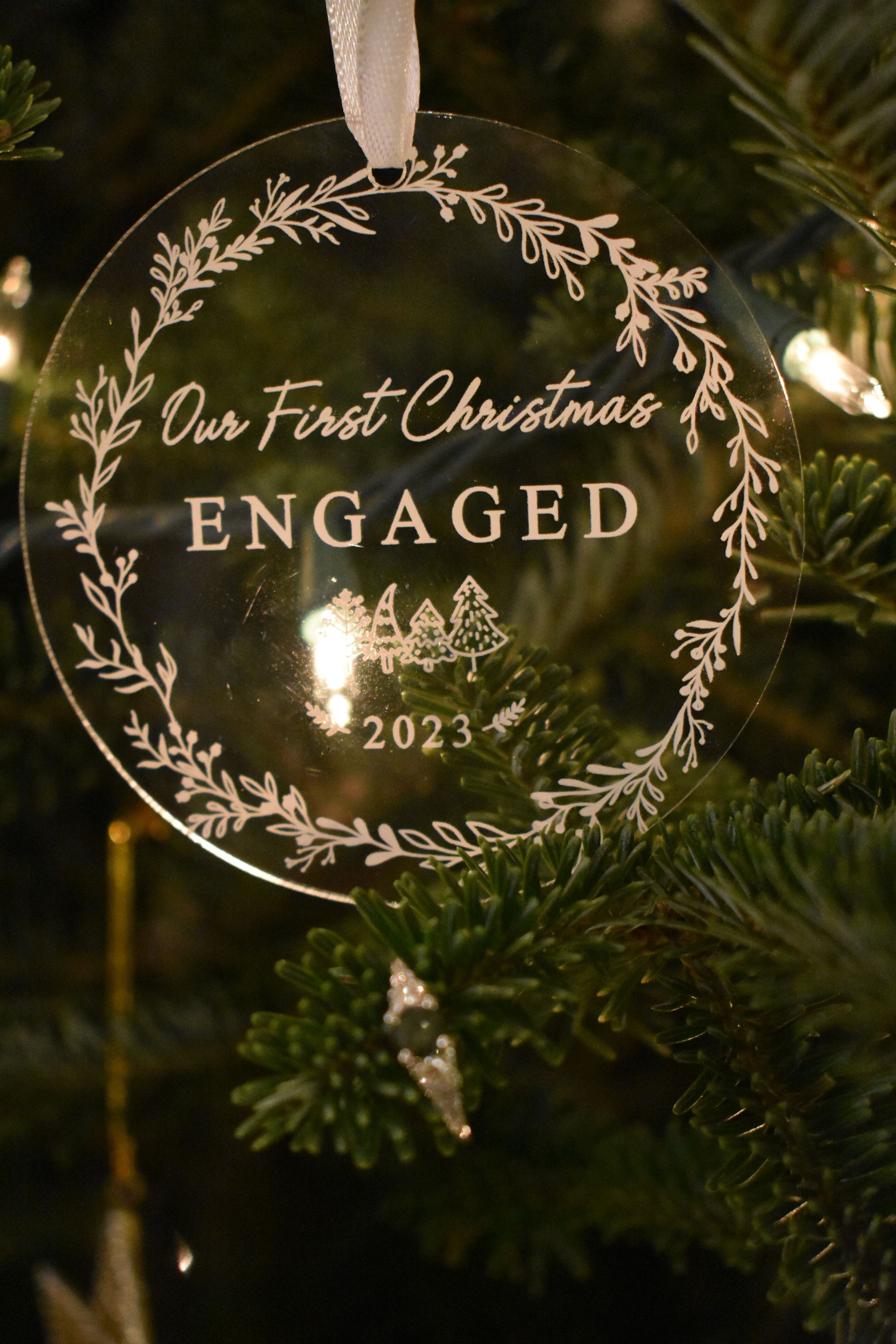 Clear glass Christmas ornament reading 'Our First Christmas Engaged 2023' with delicate floral wreath design.