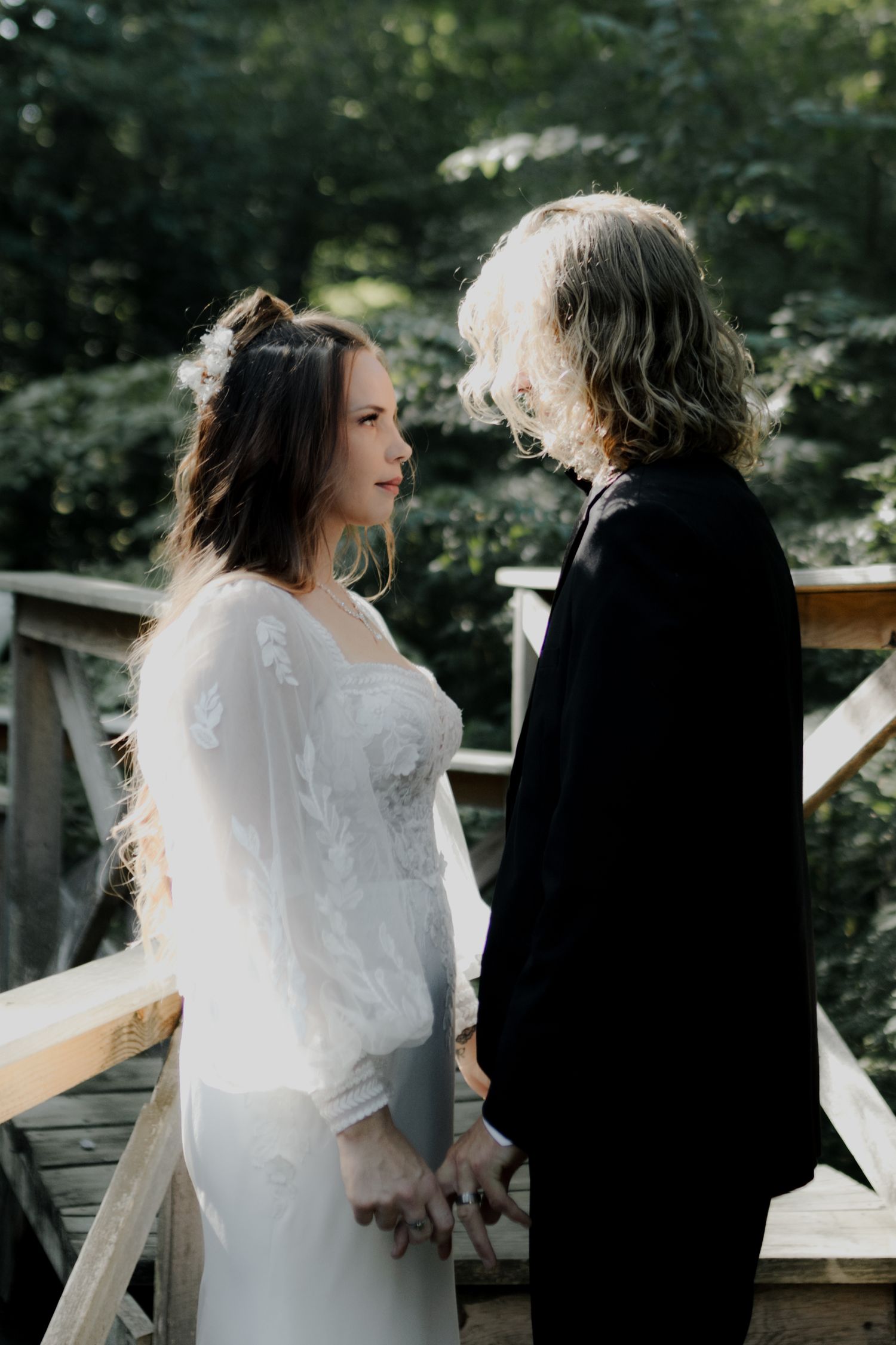 Couple in wedding attire standing on a wooden deck surrounded by trees.
