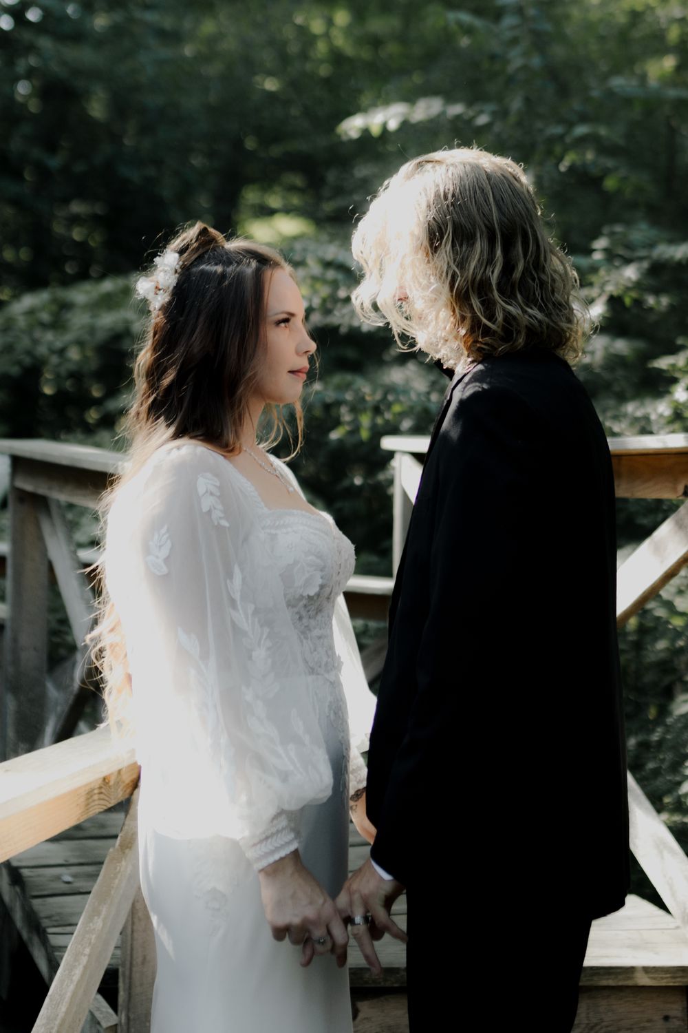 Couple in wedding attire standing on a wooden deck surrounded by trees.