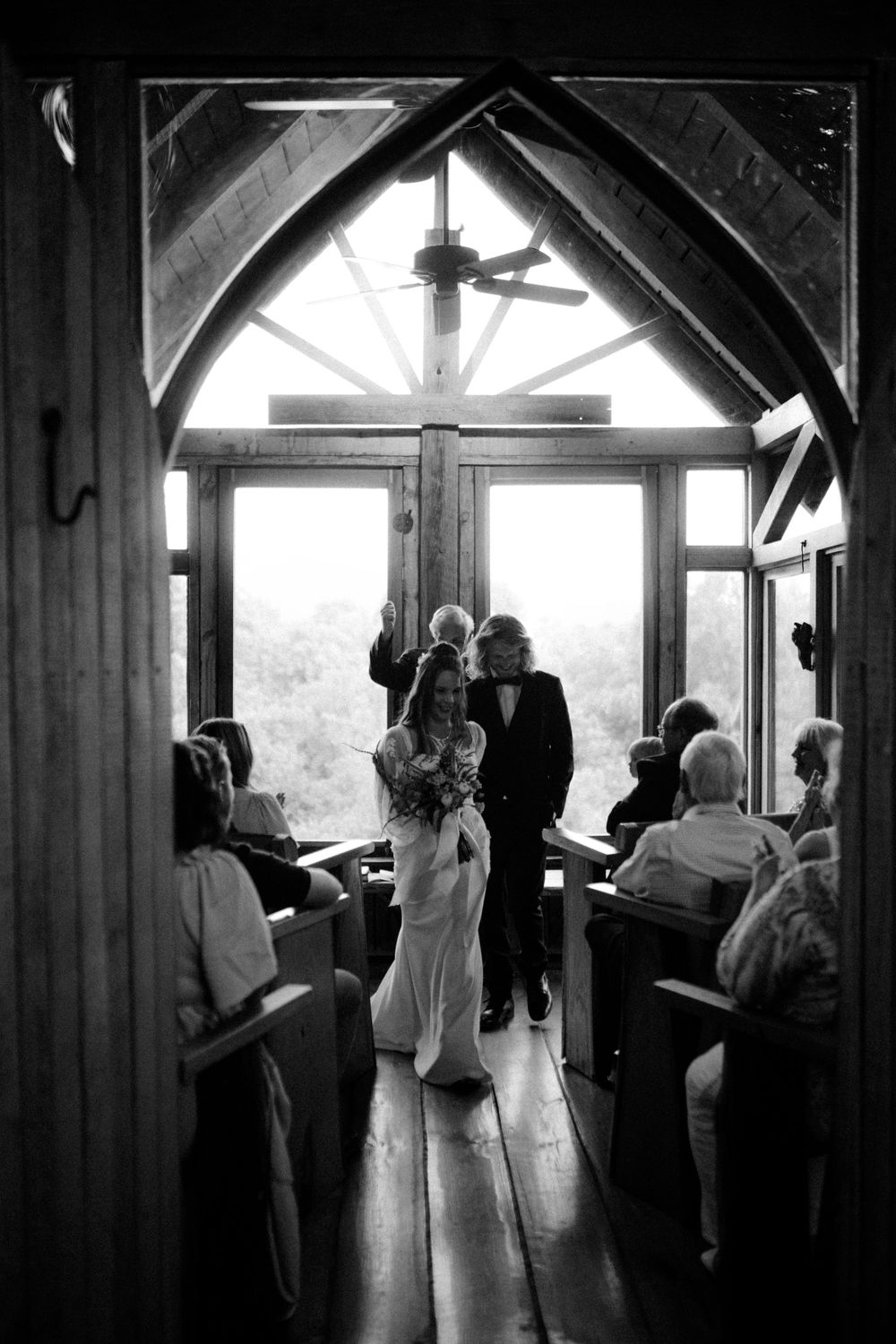 A romantic wedding moment silhouetted against bright windows in a rustic wooden chapel.