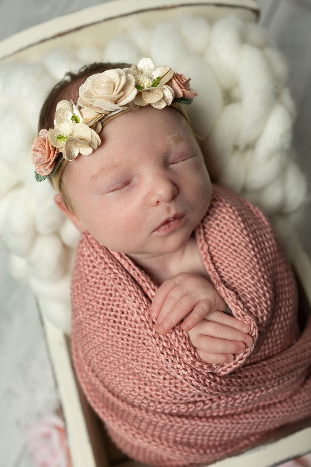 A sleeping newborn baby swaddled in a pink knit wrap wearing a delicate floral headband on white textured backdrop.