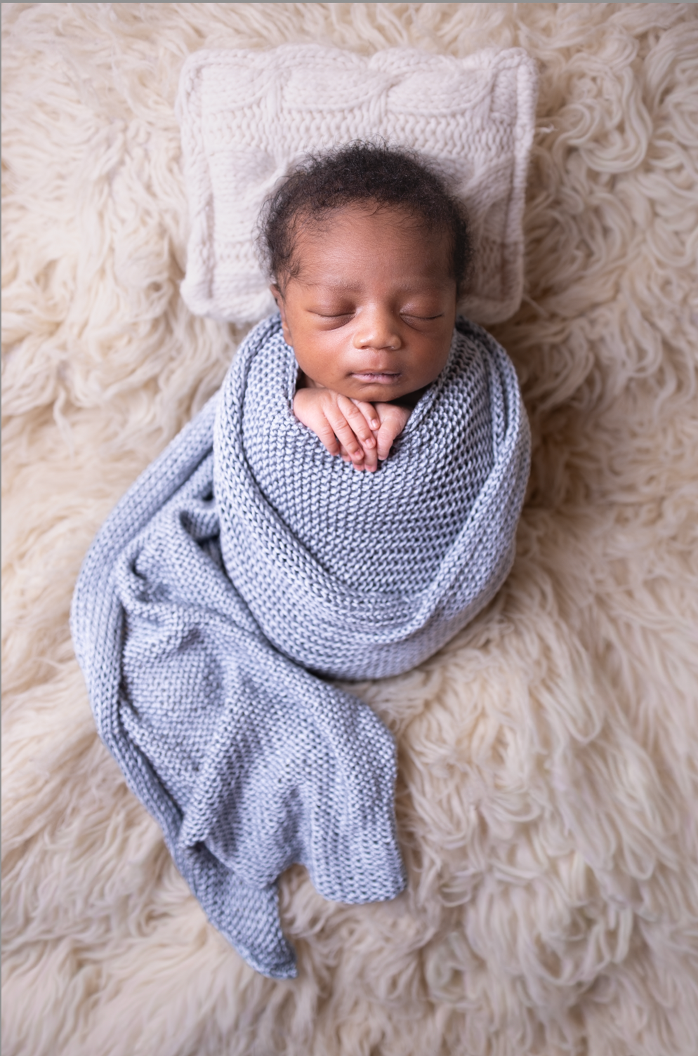 A sleeping newborn baby swaddled in a soft blue knitted blanket lies on white fluffy fur background.