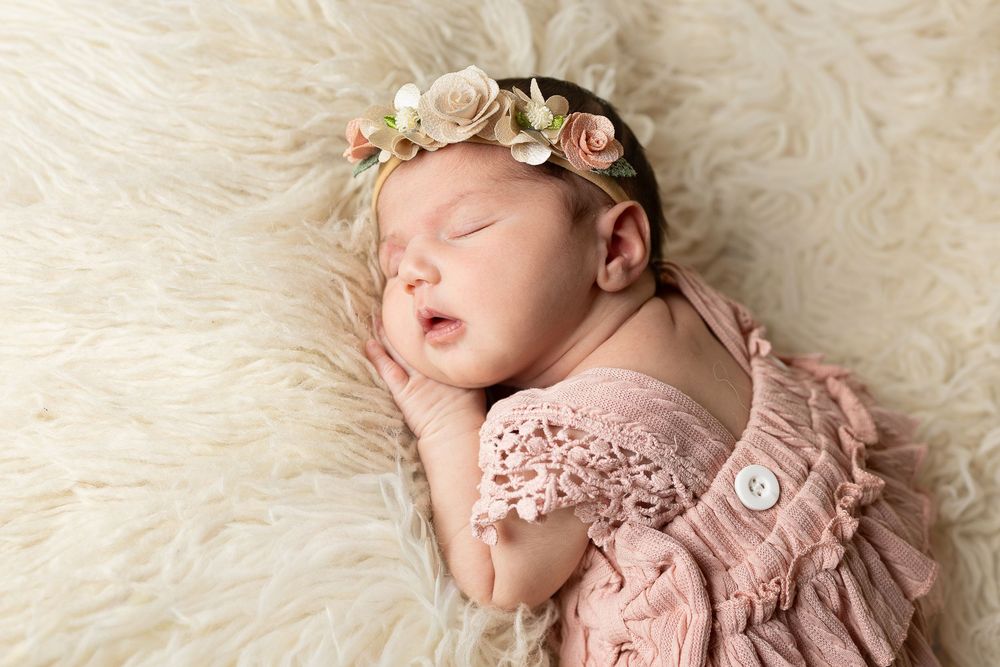 Sleeping newborn baby in pink lace dress with floral headband lying on soft cream fur backdrop.