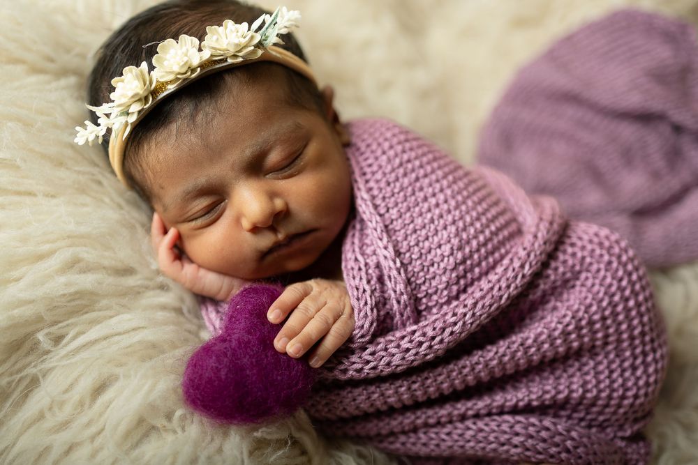Newborn baby sleeping peacefully on white fur blanket wearing purple knitted bonnet and wrap.