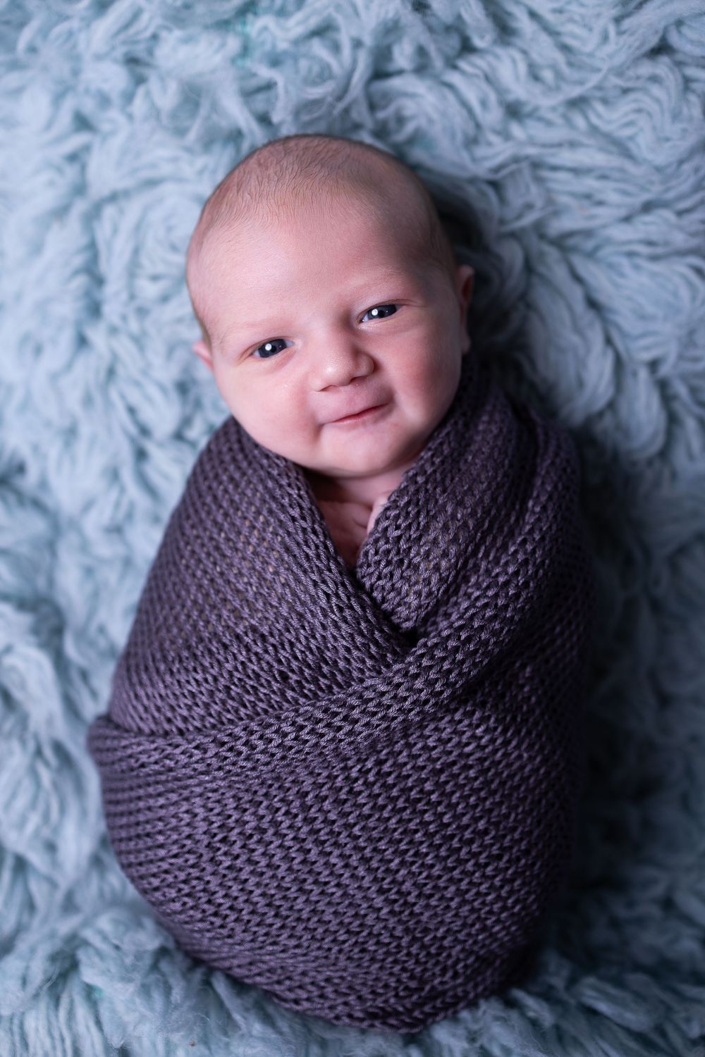 A baby wrapped in a gray knit blanket smiles while lying on a light blue textured background.