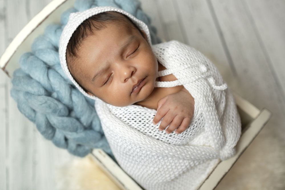 Sleeping baby swaddled in white knit blanket with blue hat resting on gray bedding.
