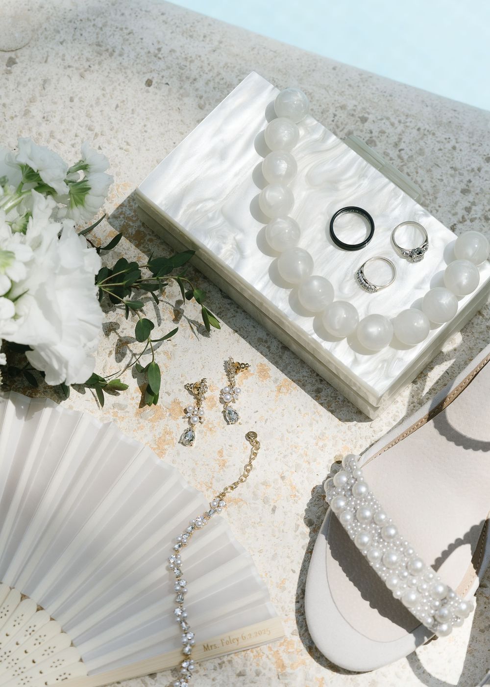 Wedding rings and delicate white paper fans displayed with pearls and floral arrangements on a textured gray surface.