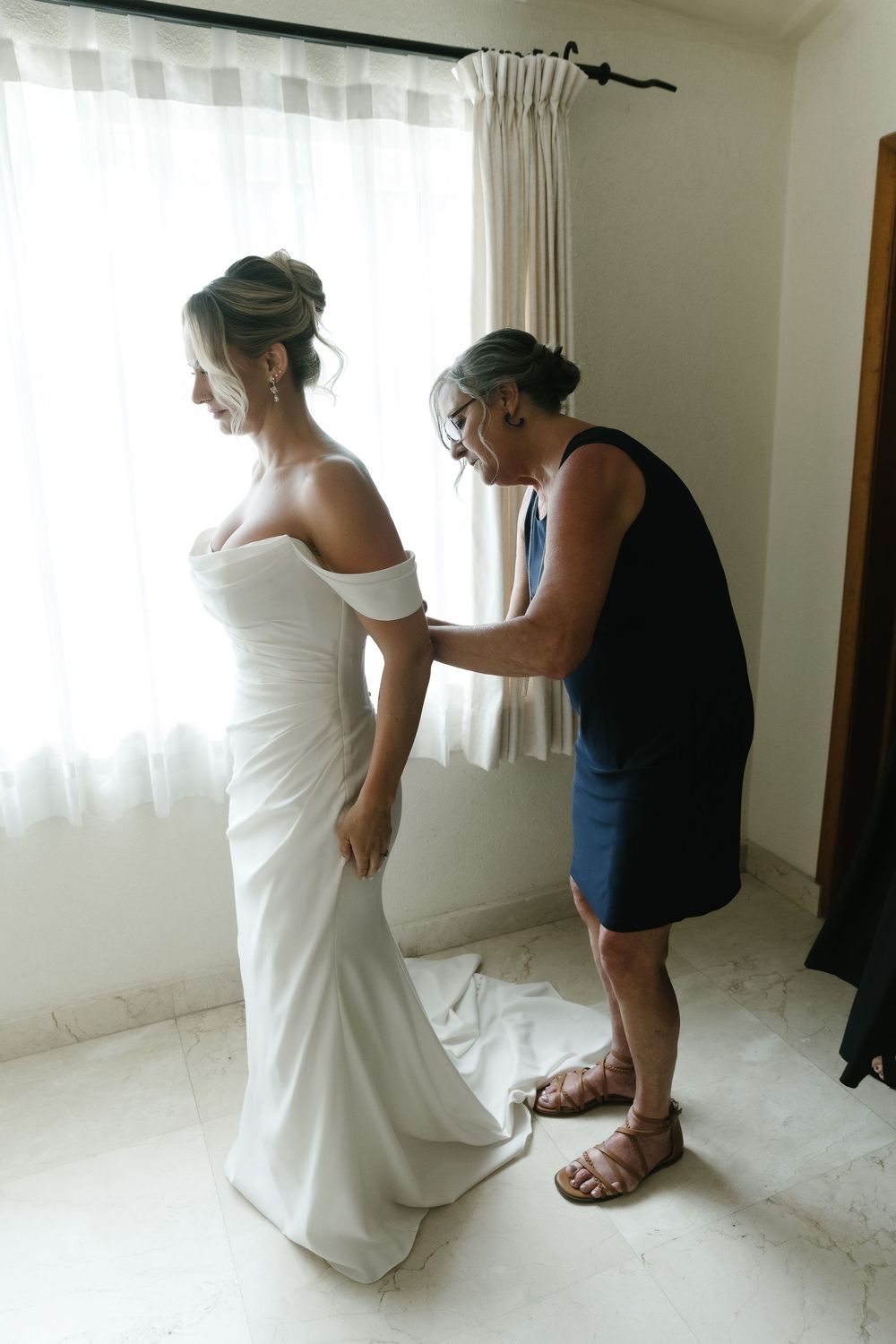 Four sequential images showing a wedding dress being fitted and adjusted in a bright white room with curtains.