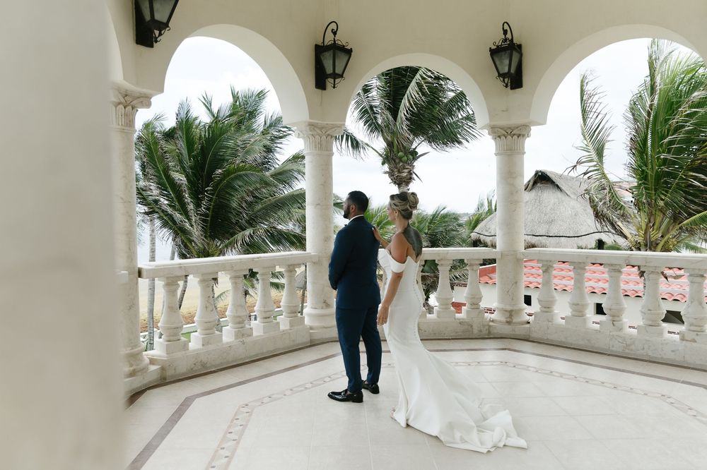 A wedding couple walks along an elegant white columned balcony with palm trees and blue sky in the background.