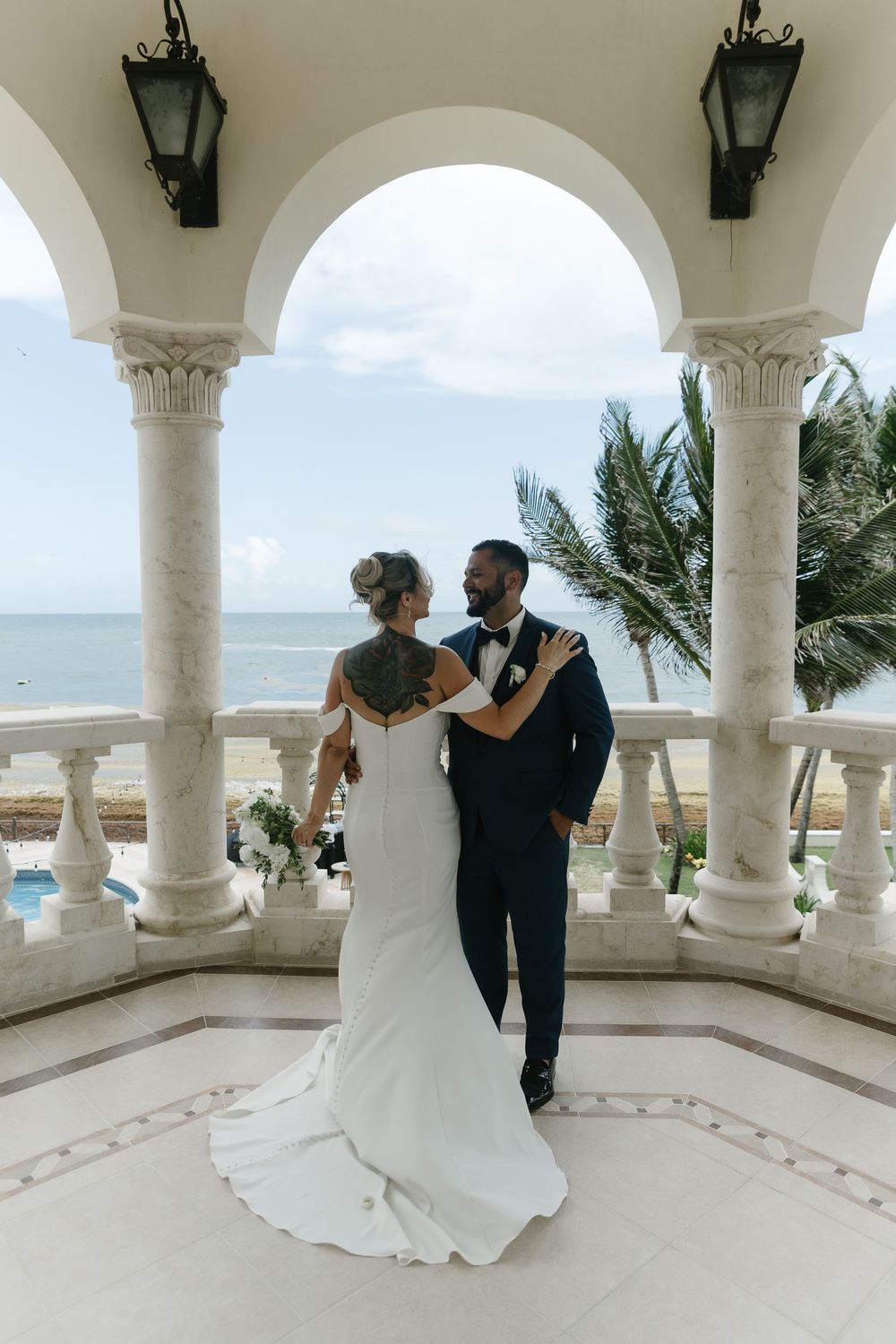 A couple shares a romantic dance moment beneath elegant white archways overlooking an ocean view at a luxury venue.