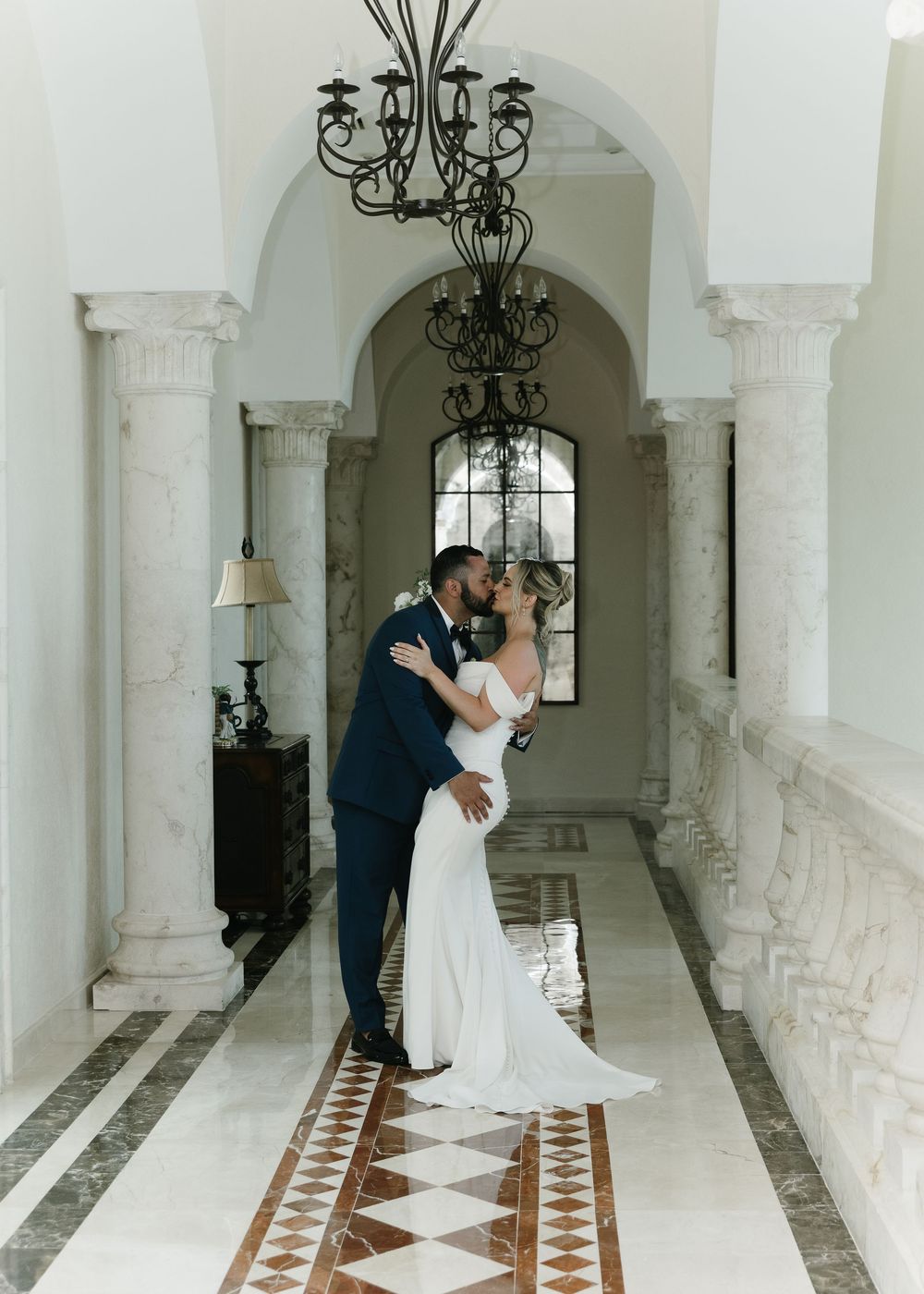 A romantic couple shares an intimate moment in an elegant white colonnade hallway with ornate chandeliers and tiled floors.