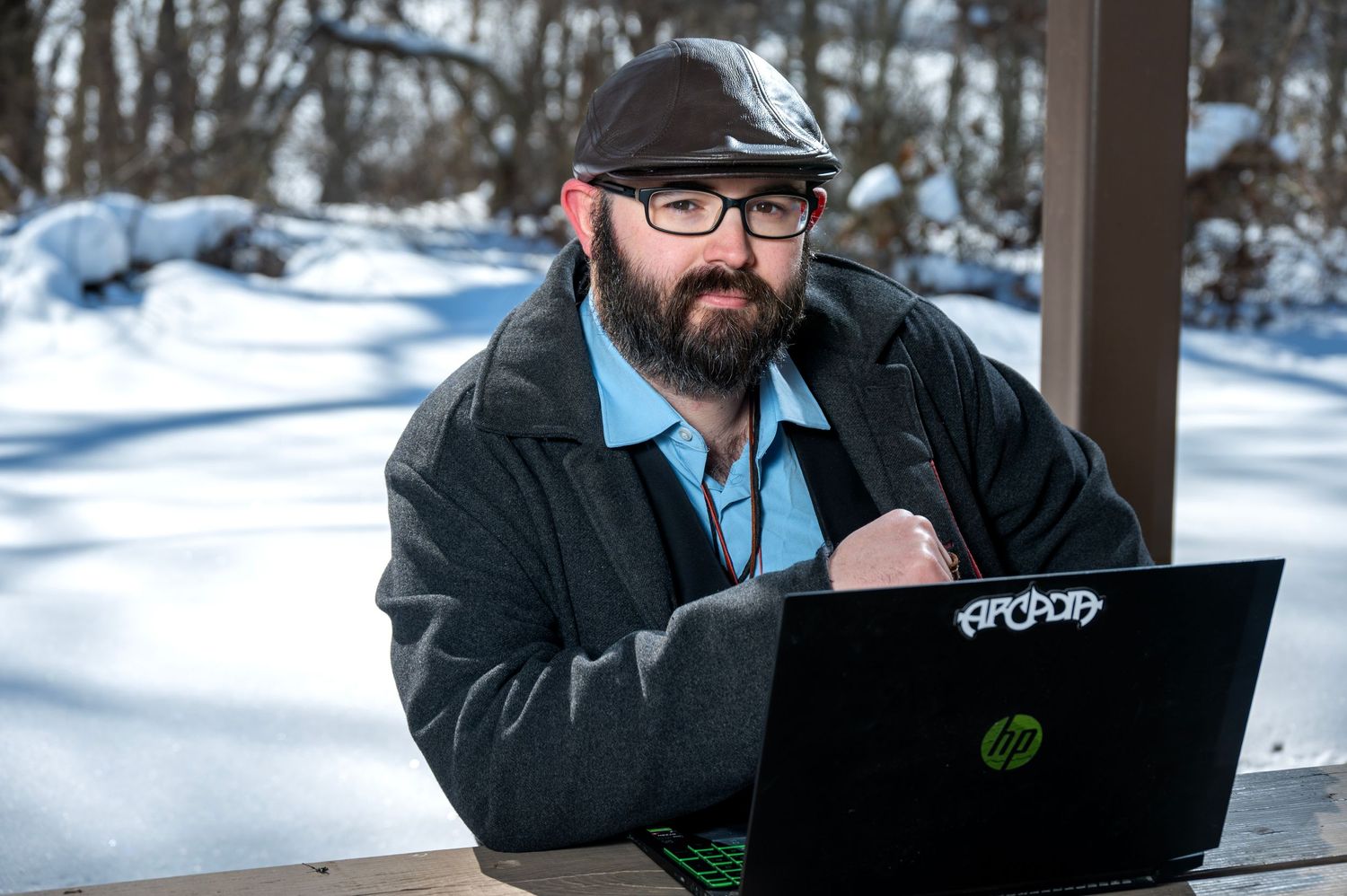Man in glasses and winter coat works on laptop outdoors in snowy setting.