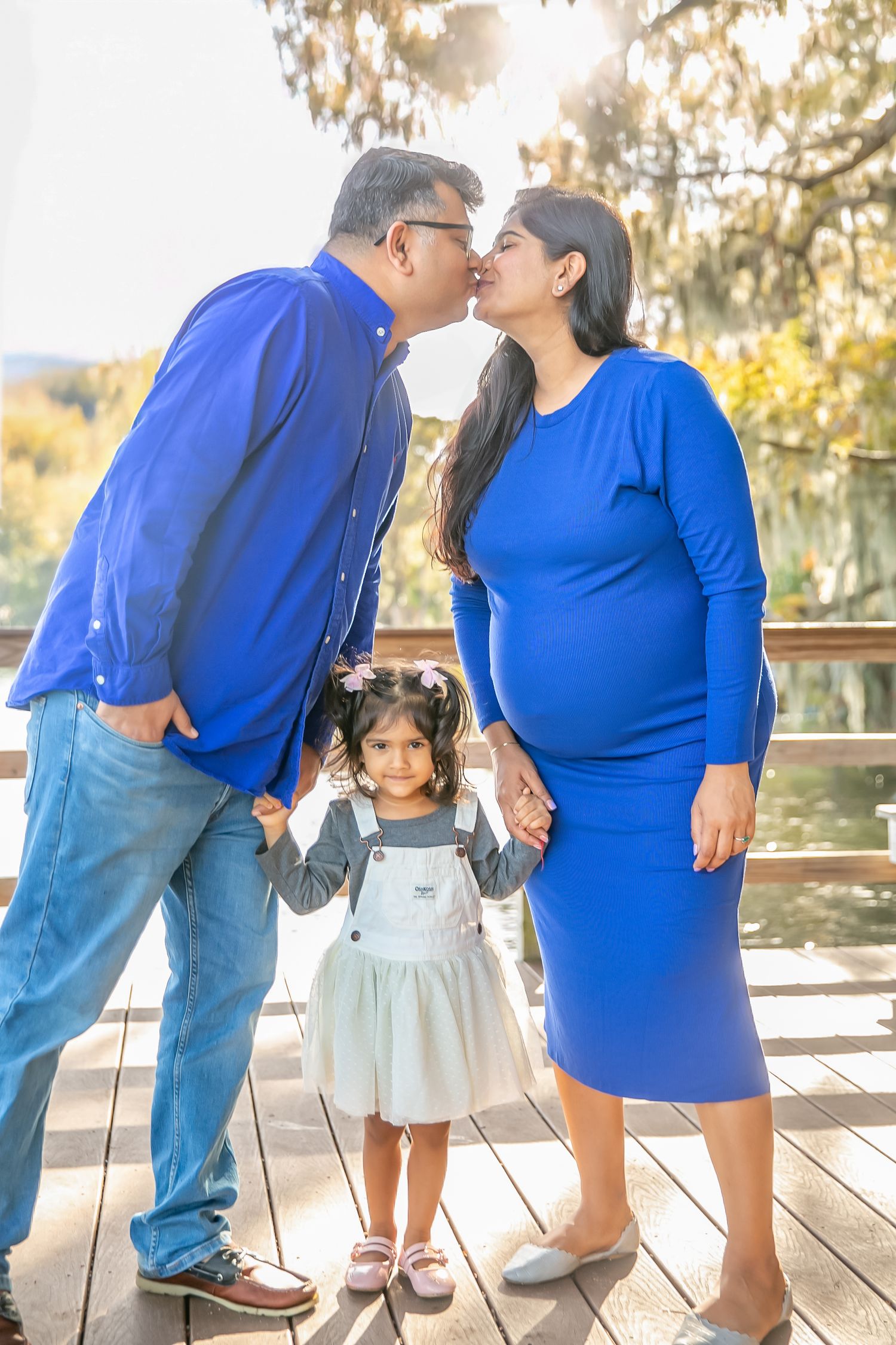 Outdoor family maternity photo in coordinating blue outfits holding white baby clothes.