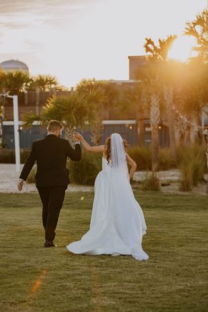 A couple walks hand in hand across a grassy lawn during golden hour at their wedding.