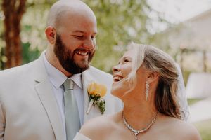 A joyful bride and groom share a laugh on their wedding day, with the groom in a light suit and yellow boutonniere.