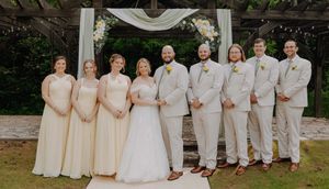 Wedding party poses outdoors in cream and beige attire before floral arch at garden ceremony venue.