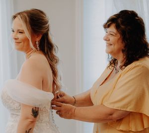 A bride in a white gown smiles as a woman in yellow helps fasten her dress buttons at a bright wedding venue.