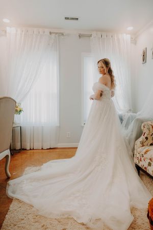 Bride in elegant off-shoulder white ballgown with long train stands by bright windows in a softly lit room.