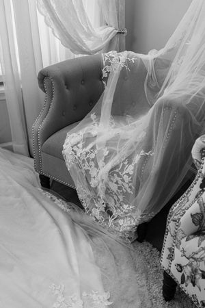 Black and white photo of an elegant lace wedding dress draped over a tufted armchair near sheer curtains.