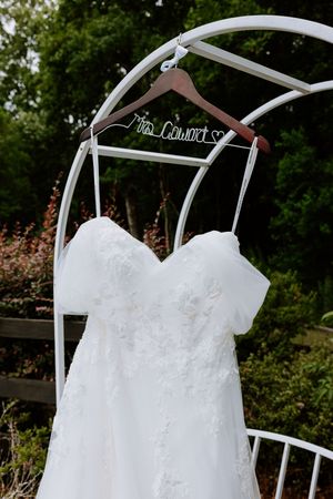 White lace wedding dress hanging on a personalized Mrs. hanger on a white garden arch surrounded by lush greenery.
