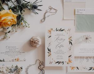 Elegant wedding invitation suite with floral design, sage green fabric swatch, jewelry, and rings on white background.