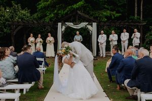 Bride and groom share first kiss at outdoor wedding ceremony with guests seated on white chairs and wedding party standing behind.