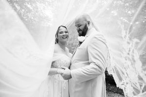 Black and white photo of a bride and groom smiling under a flowing veil on their wedding day.