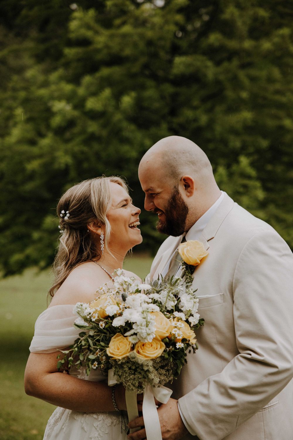 Smiling bride and groom share a joyful moment outdoors, bride holding yellow and white bouquet, groom in tan suit.