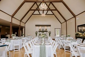 Elegant wedding reception hall with white chairs, draped tables, greenery centerpieces, and a vaulted ceiling with chandelier.