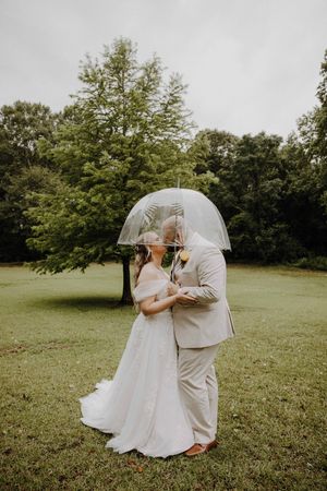 Bride and groom kissing under a clear dome umbrella on a lush green lawn during a rainy outdoor wedding.