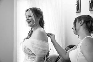 Black and white photo of a bride smiling as a bridesmaid helps fasten her elegant wedding dress near a bright window.